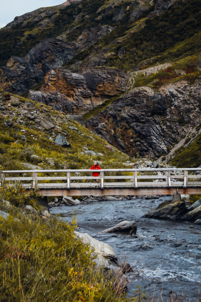 A woman in a red jacket crossing a bridge with a large rock face behind her.