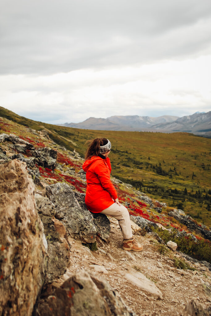 A woman in a red jacket sitting on a rock face with mountains in the background.