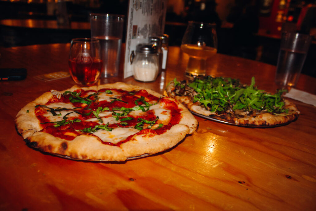 Two pizzas, one with red sauce and one with a lot of arugula, hard ciders in the background.