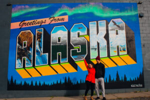 A man and a woman stand in front of a mural that says "Alaska." Each letter has a painting of nature within it.