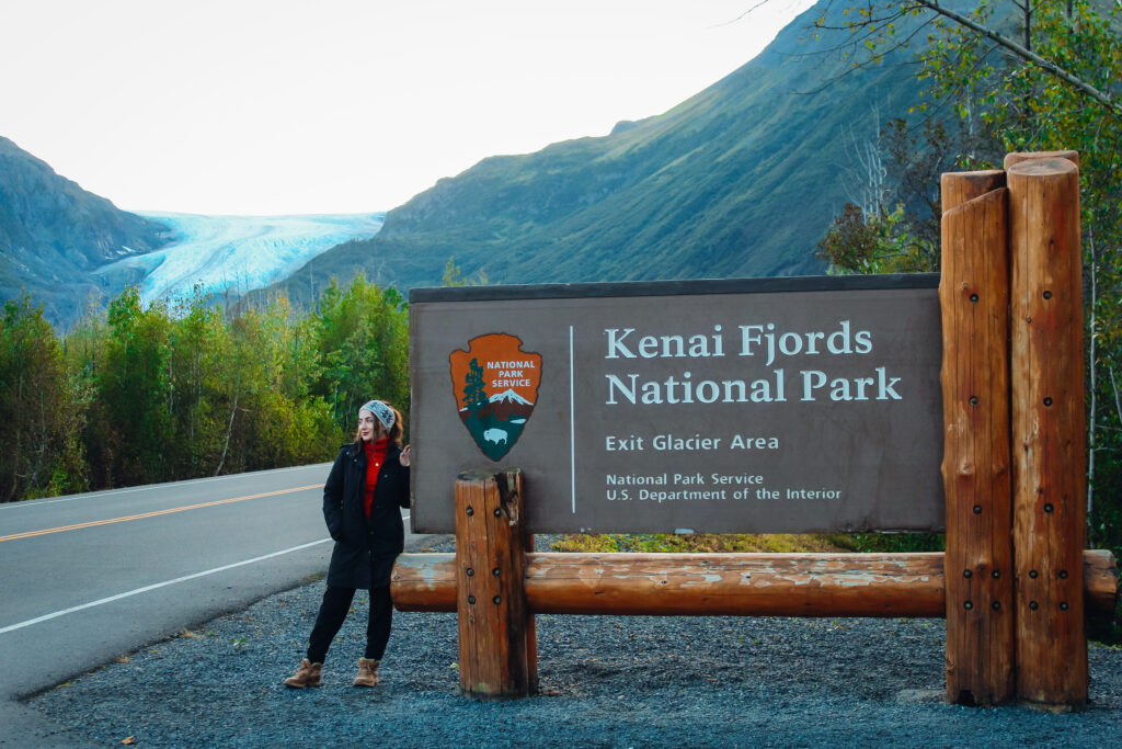 Woman standing next to Kenai Fjords National Park sign with a glacier in the background