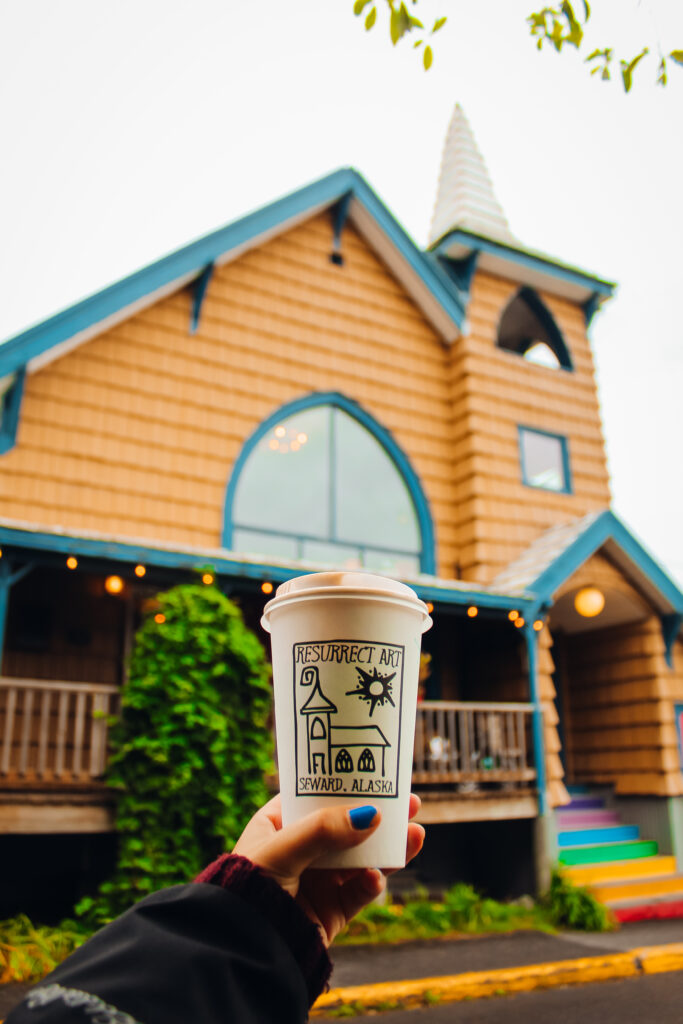 A coffee cup that says "Resurrect Art" in front of a church that is on the logo.
